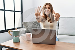 Young brunette woman using laptop at home drinking a cup of coffee showing and pointing up with fingers number six while smiling