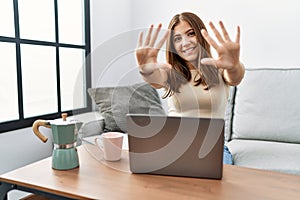 Young brunette woman using laptop at home drinking a cup of coffee showing and pointing up with fingers number ten while smiling
