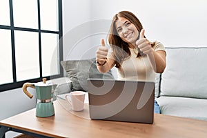 Young brunette woman using laptop at home drinking a cup of coffee success sign doing positive gesture with hand, thumbs up