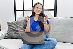 Young brunette woman using laptop at home excited for success with arms raised and eyes closed celebrating victory smiling