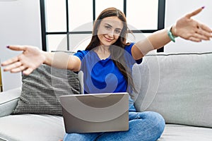 Young brunette woman using laptop at home looking at the camera smiling with open arms for hug