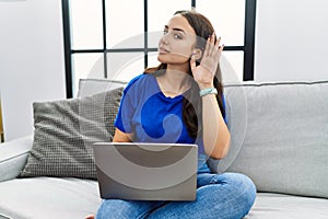 Young brunette woman using laptop at home smiling with hand over ear listening an hearing to rumor or gossip