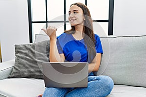Young brunette woman using laptop at home smiling with happy face looking and pointing to the side with thumb up