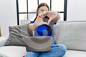 Young brunette woman using laptop at home smiling in love doing heart symbol shape with hands