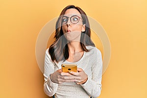 Young brunette woman using smartphone over yellow background making fish face with mouth and squinting eyes, crazy and comical