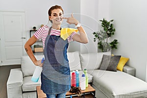 Young brunette woman wearing apron holding cleaning products at home pointing with hand finger to face and nose, smiling cheerful