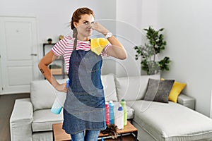 Young brunette woman wearing apron holding cleaning products at home smelling something stinky and disgusting, intolerable smell,