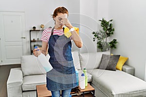 Young brunette woman wearing apron holding cleaning products at home tired rubbing nose and eyes feeling fatigue and headache