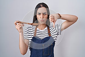 Young brunette woman wearing apron tasting food holding wooden spoon with angry face, negative sign showing dislike with thumbs