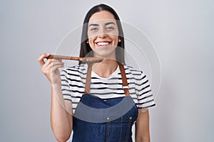 Young brunette woman wearing apron tasting food holding wooden spoon looking positive and happy standing and smiling with a