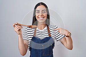 Young brunette woman wearing apron tasting food holding wooden spoon pointing finger to one self smiling happy and proud