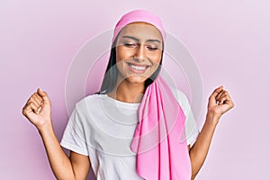 Young brunette woman wearing breast cancer support pink scarf very happy and excited doing winner gesture with arms raised,