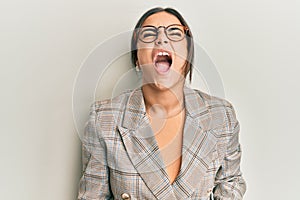 Young brunette woman wearing business jacket and glasses angry and mad screaming frustrated and furious, shouting with anger