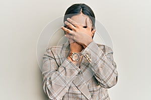 Young brunette woman wearing business jacket and glasses covering eyes and mouth with hands, surprised and shocked