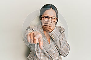 Young brunette woman wearing business jacket and glasses laughing at you, pointing finger to the camera with hand over mouth,