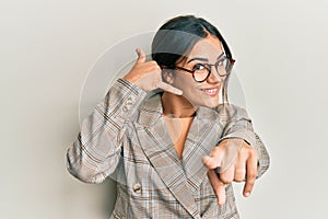 Young brunette woman wearing business jacket and glasses smiling doing talking on the telephone gesture and pointing to you