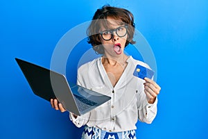 Young brunette woman wearing business style holding laptop and credit card in shock face, looking skeptical and sarcastic,