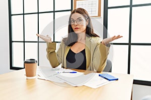 Young brunette woman wearing business style sitting on desk at office clueless and confused expression with arms and hands raised