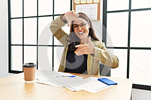 Young brunette woman wearing business style sitting on desk at office smiling making frame with hands and fingers with happy face