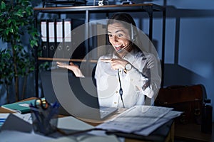 Young brunette woman wearing call center agent headset working late at night amazed and smiling to the camera while presenting