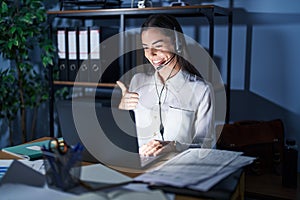 Young brunette woman wearing call center agent headset working late at night doing happy thumbs up gesture with hand