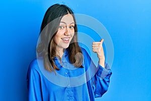 Young brunette woman wearing casual blue shirt smiling with happy face looking and pointing to the side with thumb up