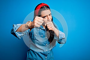 Young brunette woman wearing casual denim shirt over blue isolated background Punching fist to fight, aggressive and angry attack,