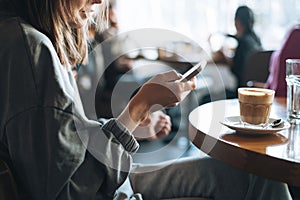 Young brunette woman wearing casual longsleeve with mobile phone in hands with cup of coffee sitting in cafe, city life