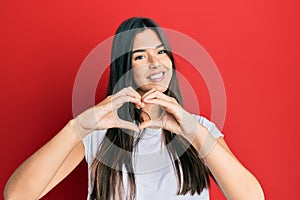 Young brunette woman wearing casual white tshirt over red background smiling in love doing heart symbol shape with hands