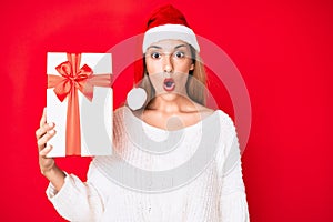 Young brunette woman wearing christmas hat and holding a gift scared and amazed with open mouth for surprise, disbelief face