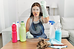Young brunette woman wearing cleaner apron and gloves cleaning at home angry and mad raising fist frustrated and furious while photo