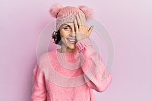 Young brunette woman wearing cute wool cap covering one eye with hand, confident smile on face and surprise emotion