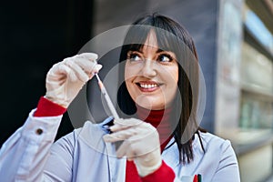 Young brunette woman wearing doctor uniform holding syringe and vaccine at the city