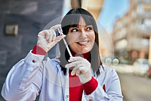 Young brunette woman wearing doctor uniform holding syringe and vaccine at the city