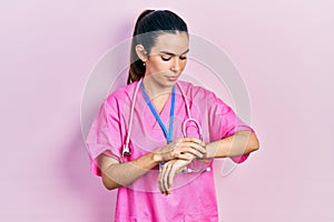 Young brunette woman wearing doctor uniform and stethoscope checking the time on wrist watch, relaxed and confident