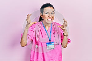 Young brunette woman wearing doctor uniform and stethoscope shouting frustrated with rage, hands trying to strangle, yelling mad