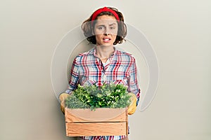 Young brunette woman wearing gardener clothes holding wooden plant pot clueless and confused expression