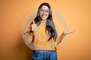Young brunette woman wearing glasses and casual sweater over yellow isolated background Showing palm hand and doing ok gesture