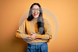 Young brunette woman wearing glasses and casual sweater over yellow isolated background smiling and laughing hard out loud because