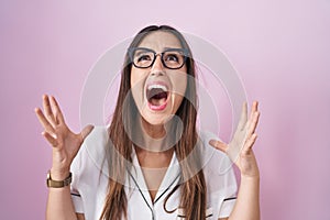 Young brunette woman wearing glasses standing over pink background crazy and mad shouting and yelling with aggressive expression