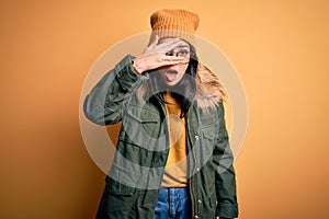 Young brunette woman wearing glasses and winter coat with hat over yellow isolated background peeking in shock covering face and