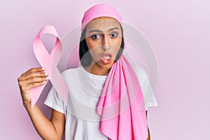 Young brunette woman wearing head scarf and holding pink cancer ribbon scared and amazed with open mouth for surprise, disbelief