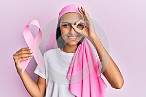 Young brunette woman wearing head scarf and holding pink cancer ribbon smiling happy doing ok sign with hand on eye looking