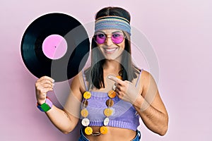 Young brunette woman wearing hippie style holding vinyl disc smiling happy pointing with hand and finger