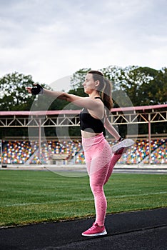 Young brunette woman, wearing pink leggings, sneakers, black top and fitness gloves, training on city stadium in morning.