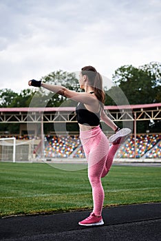 Young brunette woman, wearing pink leggings, sneakers, black top and fitness gloves, training on city stadium in morning.