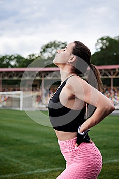 Young brunette woman, wearing pink leggings, sneakers, black top and fitness gloves, training on city stadium in summer.