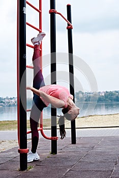 Young brunette woman, wearing pink top and blue leggings, hanging on wall bars doing twine outside by city lake. Slim fit