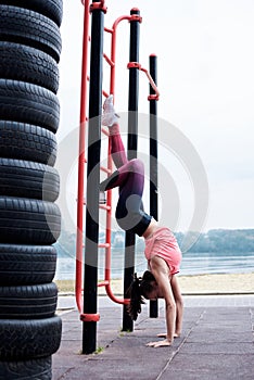 Young brunette woman, wearing pink top and blue leggings, hanging on wall bars outside by city lake. Slim fit sportswoman, doing