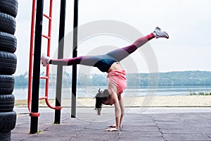 Young brunette woman, wearing pink top and blue leggings, standing on arms by wall bars, doing twine by city lake. Slim fit
